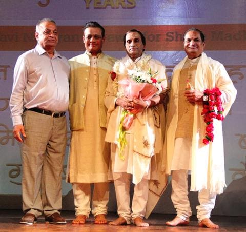 Four men on stage, one holding a bouquet, another a garland
